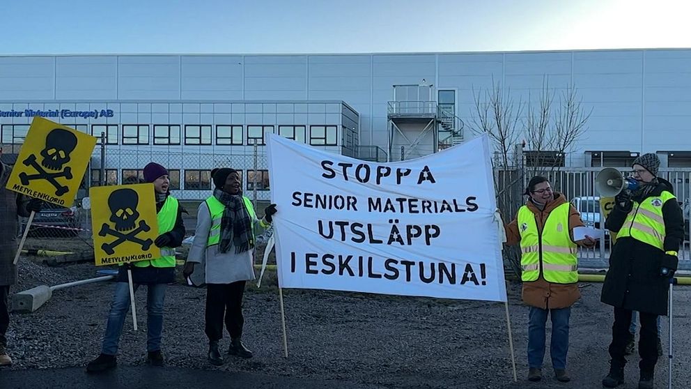 Grupp människor i gula reflexvästar håller skyltar och en banderoll framför en industribyggnad som tillhör Senior Material (Europe) AB. Banderollen har texten ”STOPPA SENIOR MATERIALS UTSLÄPP I ESKILSTUNA” och uppmanar till åtgärder mot miljöutsläpp. Några skyltar visar bilder av dödskallar för att varna för fara.