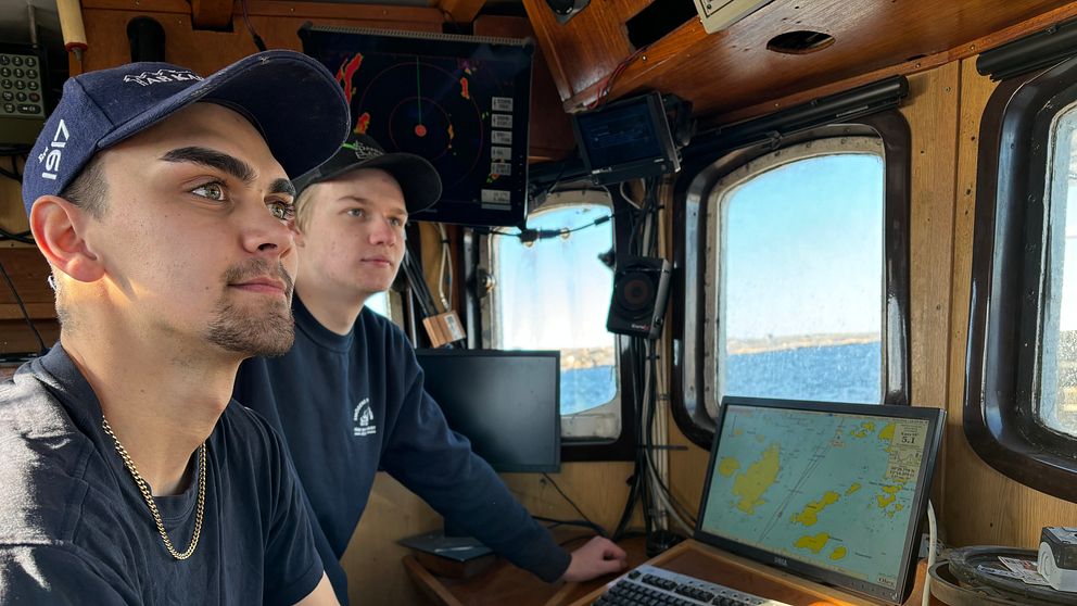 Två personer navigerar i en skeppshytt med havsutsikt och radarutrustning.