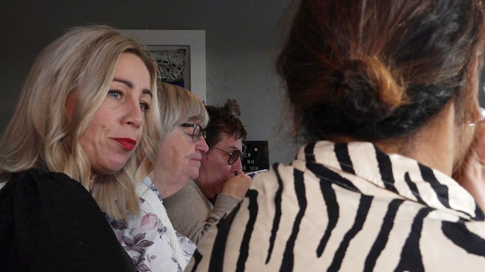 Flera personer sitter samlade i ett rum, där två av dem är delvis skymda. En kvinna till vänster har långt, ljust hår och bär en mörk tröja. . I förgrunden syns en kvinna med mörkt hår, som har på sig en randig klädsel. En delvis skymd person i bakgrunden dricker ur en kopp.