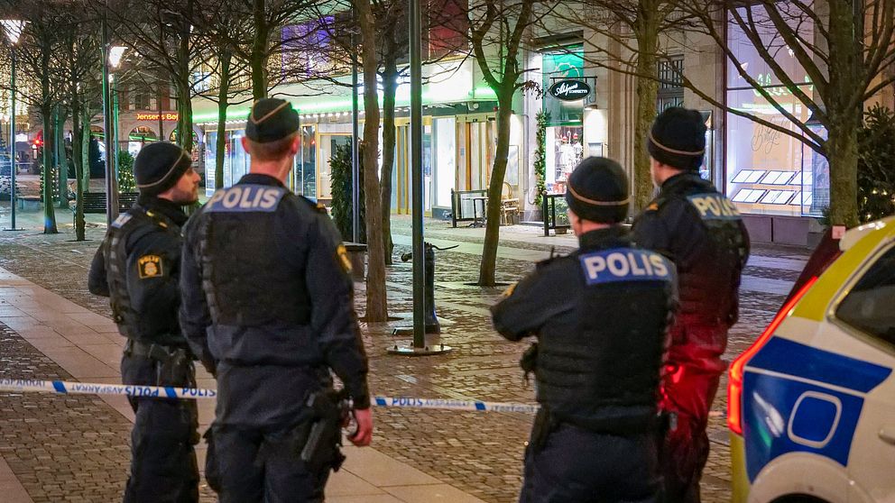 Fyra poliser står på en trottoar under natten, observerande mot en avspärrning med polisband. I bakgrunden syns en butik som är belyst med färgglad belysning, och en polisbil är parkerad i närheten. Bilden är tagen i centrala Malmö.