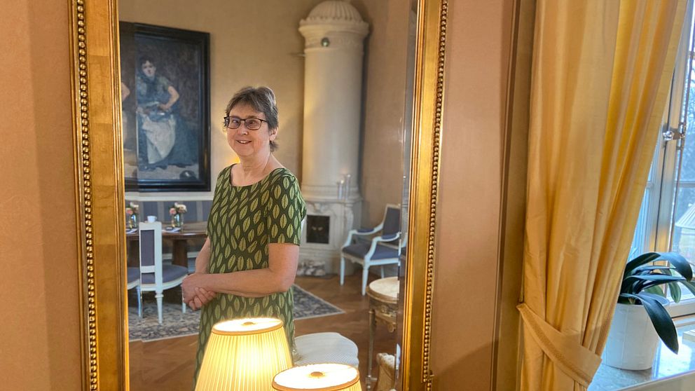 Landshövding Brittis Benzler står i sitt hem och tittar in i kameran via en spegel med förgylld ram.