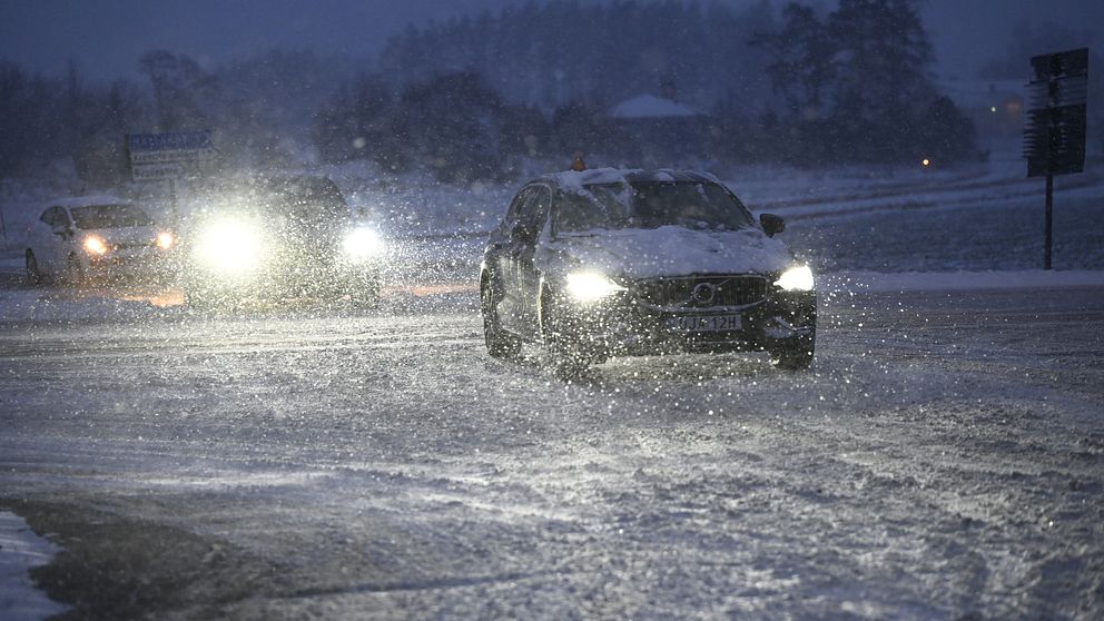 En bil kör i snöigt och dimmigt väder, med snöflingor som virvlar runt. Två andra bilar syns i bakgrunden med sina strålkastare tända. Känsla av en kall vinterkväll.