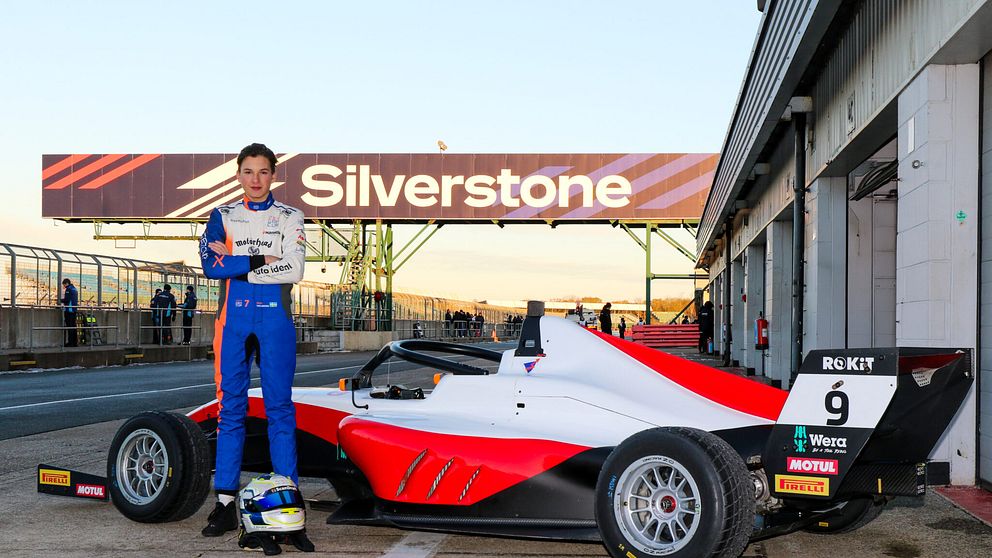 En ung förare står bredvid en racingbil på Silverstone-banan, med en stor skylt som visar banans namn i bakgrunden. Föraren är klädd i en blå och orange racingdräkt och håller en hjälm i handen. Racingbilen är vit med röda och svarta detaljer och har nummer 9 på sidan.