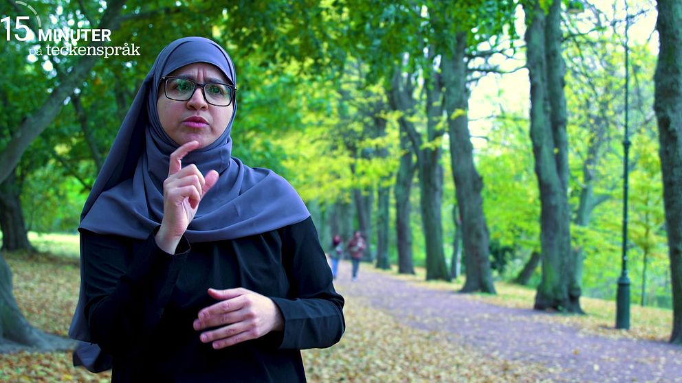 Fatima Eisa har svarta kläder och en grå sjal. Hon står i en park med gröna träd och fallna löv runtomkring. I bakgrunden syns en stig där en annan person promenerar. Hon tecknar ordet muslim.