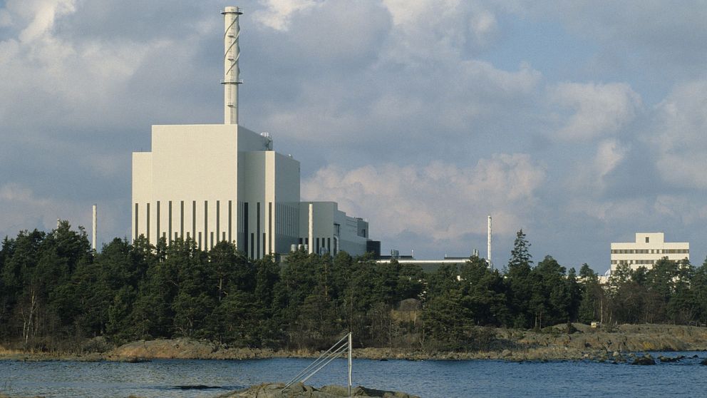 Kärnkraftverk vid kusten, omgiven av vatten och skog under molnig himmel.