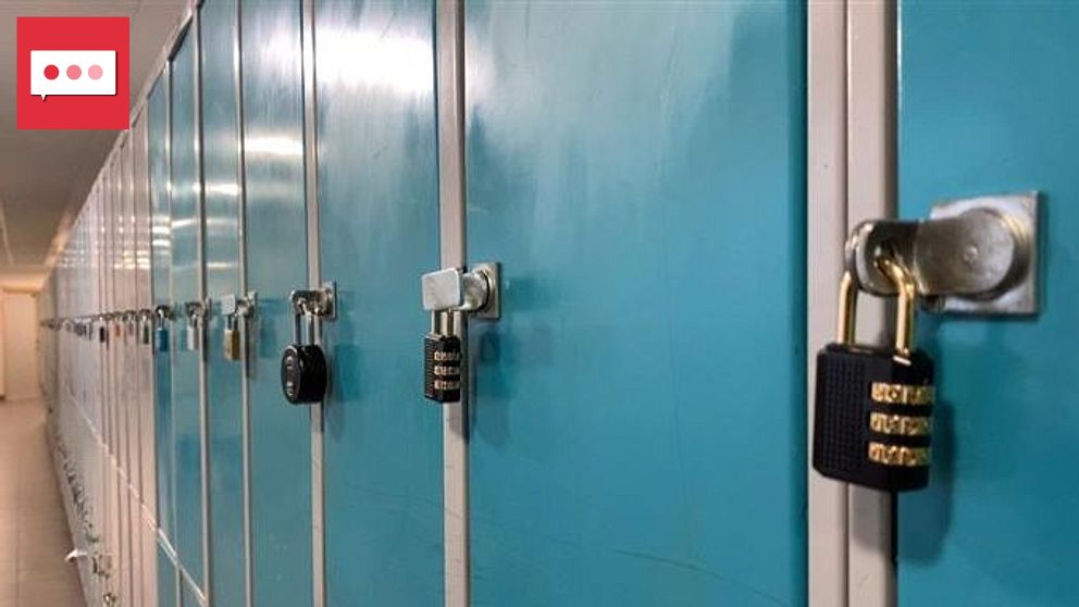 Rader av blåa skåp med hänglås på en skolkorridor, en person går längst bort i bakgrunden.