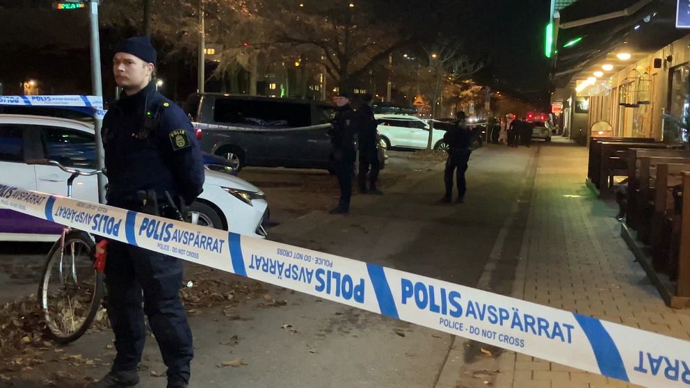 Poliser står vid en avspärrning på en gata, omgiven av flera fordon. Bredvid en cykel är avspärrningsbandet tydligt synligt med texten ”POLIS AVSPÄRRAT”. I bakgrunden syns fler poliser som arbetar i området, under en mörk kväll.