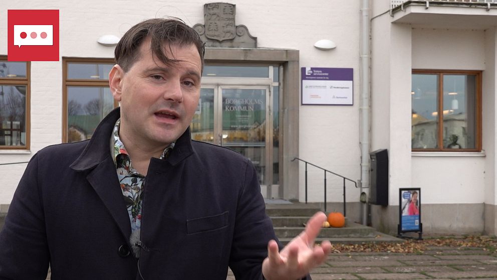 Personen, Joel Schäfer (S), ordförande i socialnämnden i Borgholms kommun gestikulerar och talar framför en byggnad med en dörr som visar namnet Borgholms kommun. I bakgrunden syns en orange pumpa och fönster med synligt ljus. Det är en grå och mulen dag.
