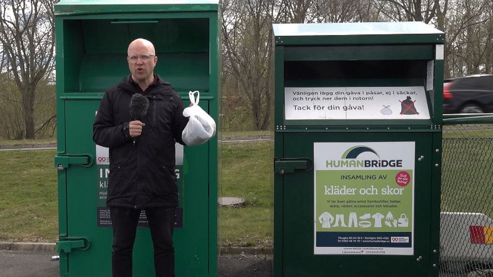 En reporter håller upp en plastpåse med kläder i. Han står framför gröna insamlingsboxar.