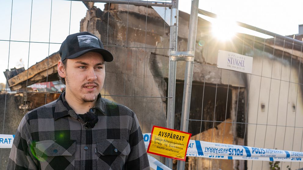 Alvar Claesson står framför en avspärrning vid en brandskadad byggnad. I bakgrunden syns resterna av byggnaden med tydliga skador, och en solstråle bryter genom den krisande strukturen. En röd och gul avspärrningsskylt är placerad i bildens nedre del.
