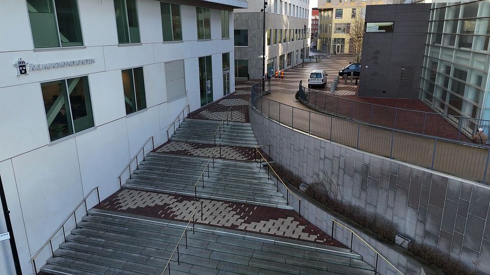 Trappor som leder ner från en byggnad med arkitektonisk design, omgivna av en innegård och andra byggnader. En bil är parkerad i bakgrunden.
