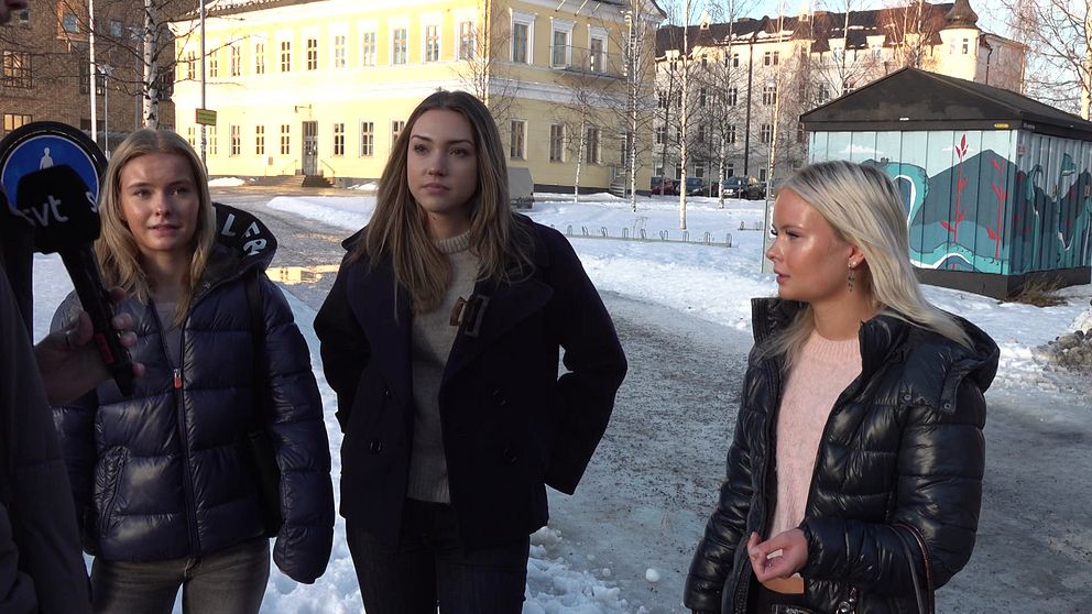 Tre tjejer står på en snötäckt gata, klädda i vinterjackor, med en byggnad i bakgrunden och en färgglad muralmålning.