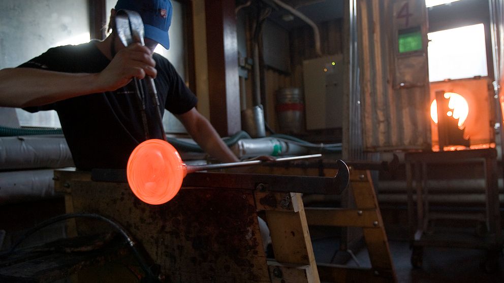 Glasblåsare formar glödande glas i en verkstad med ugn i bakgrunden.
