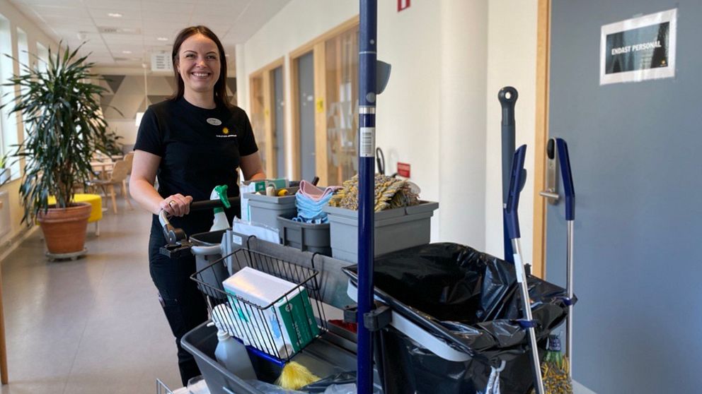 En lokalvårdare skjuter en städvagn med rengöringsprodukter och redskap i en korridor. Till vänster syns en stor krukväxt och till höger en dörr med skylten ”Endast personal”.