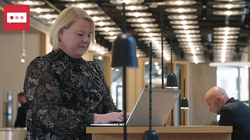 En person står vid en stående arbetsstation och arbetar på en laptop i en modern, ljus arbetsmiljö. I bakgrunden syns en kollega sittande vid ett bord. Rummet är väl upplyst med moderna lampor och har trämaterialinslag.