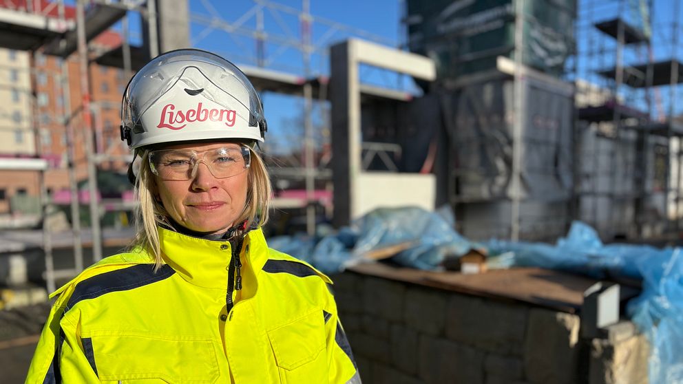 En person iklädd en gul reflexjacka och skyddshjälm står på en byggarbetsplats, omgiven av byggnadsställningar och staplade byggmaterial.