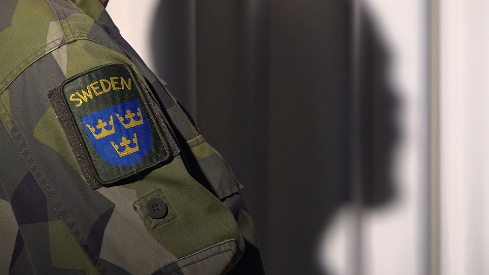 Närbild på ärmen på en person i militärkläder, skuggan av honom syns på väggen bakom.