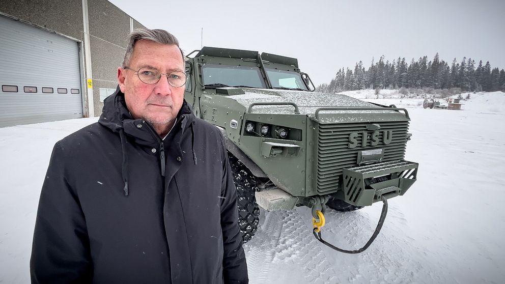 En person står i snöigt landskap framför en grön militärfordon med en robust design. Fordonet har stora däck och en kraftig framgrill, och i bakgrunden syns en byggnad och snöklädda träd.