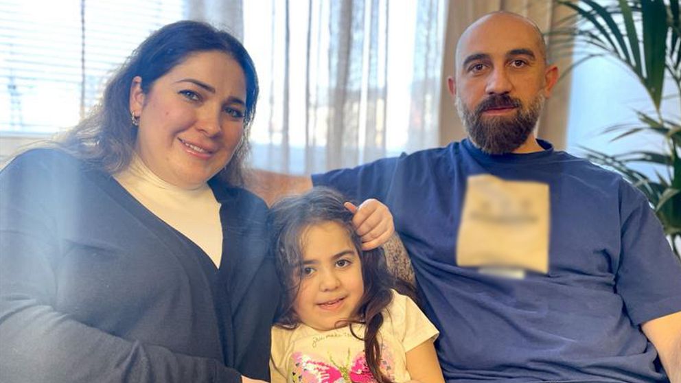 En familj sitter i ett vardagsrum med ljuset strömmande genom fönstret bakom dem. En vuxen kvinna och man sitter på varsin sida om ett barn, där mannen håller ena armen runt barnet.