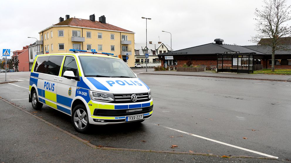 En polisbil står parkerad på en tom gata i en stadsmiljö med bostadshus och en mindre byggnad i bakgrunden under en molnig himmel.