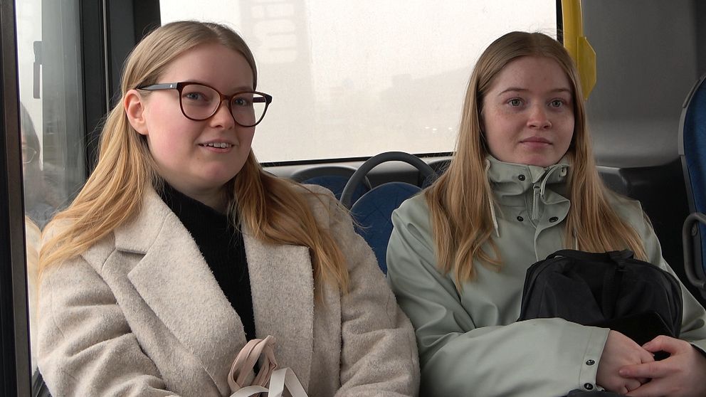 Två tjejer med långt hår sitter bredvid varann på en buss.