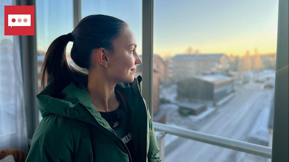 En person står vid ett fönster och blickar ut över en vinterlandskap med snöklädda gator och hus i bakgrunden. Morgonsolen lyser svagt över horisonten, vilket ger en lugn. Personen bär en grön jacka och har en avslappnad hållning.