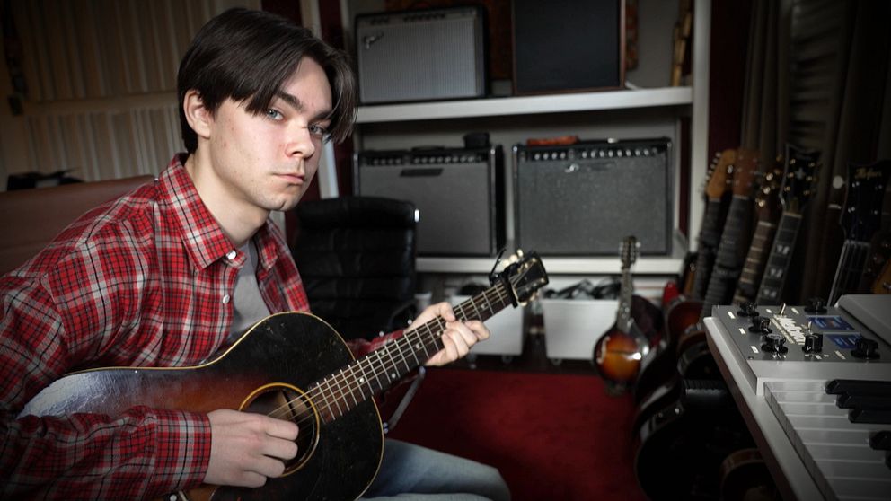 Person i röd skjorta spelar gitarr omgiven av förstärkare och musikinstrument.
