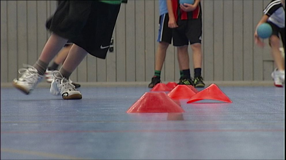 Aktiva barn som spelar inomhus, rör sig kring orange koner på ett gymnastikgolv. Fokus ligger på benen som springer mot konerna, medan andra barn syns i bakgrunden som deltar i aktiviteten. Det handlar om att fler barn och unga i Västerbotten får fysisk aktivitet på recept.