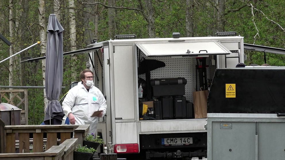 Man iklädd vit overall och munskydd går vid polisens bilar.