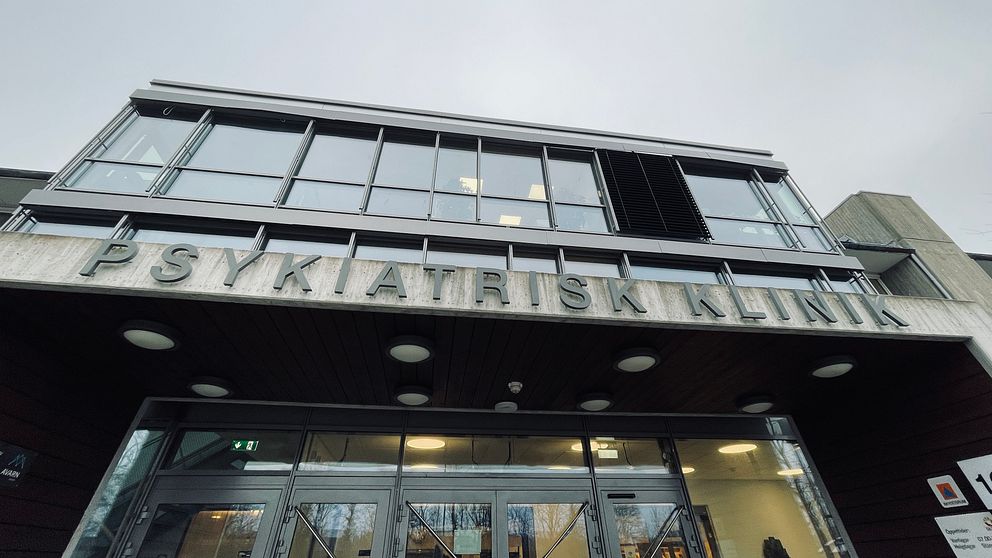 Ingången till en psykiatrisk klinik med en modern fasad. Över huvuddörrarna finns en stor skylt med texten ”PSYKIATRISK KLINIK”. Byggnaden har stora fönster som ger en ljus atmosfär. Dagen är mulen.