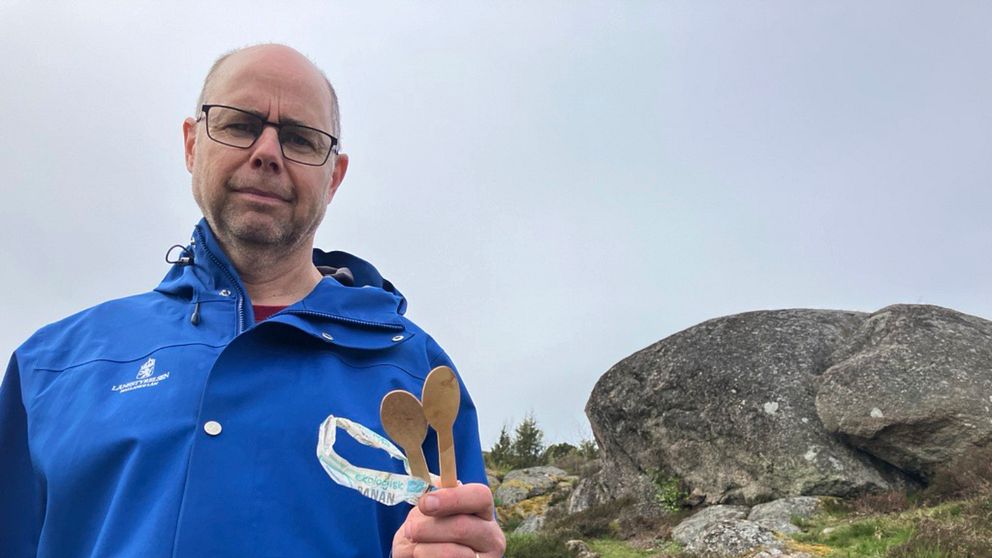 Mikael Stenström reservatförvaltare står med två träskedar och en plastbit framför en stor sten i ett naturreservat