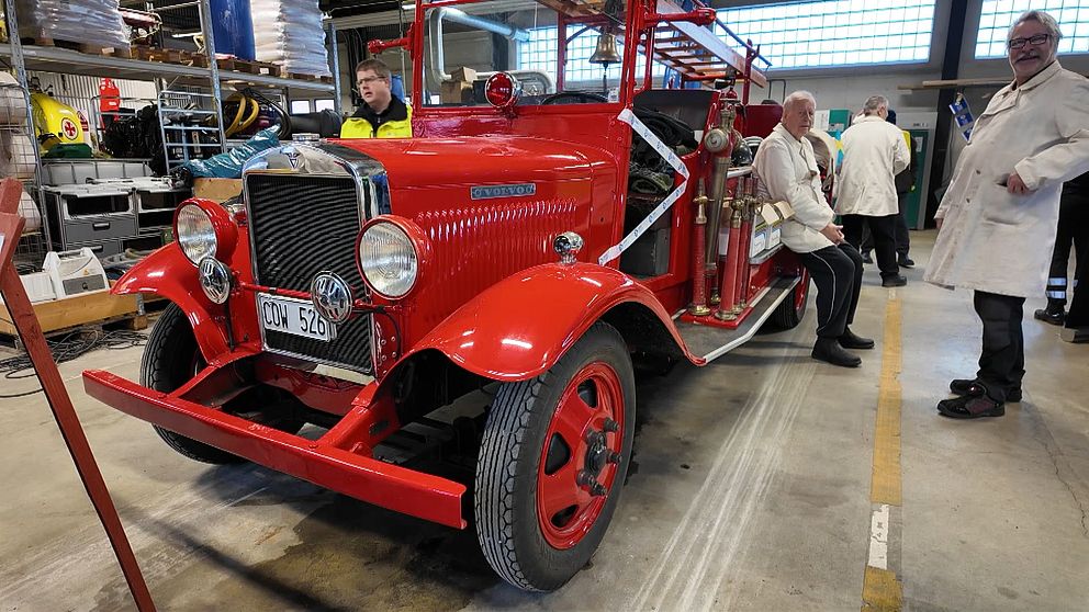 En gammal röd brandbil står inomhus i en verkstadsmiljö, omgiven av personer i arbetskläder. Brandbilen har runda strålkastare och svarta däck med röda fälgar.