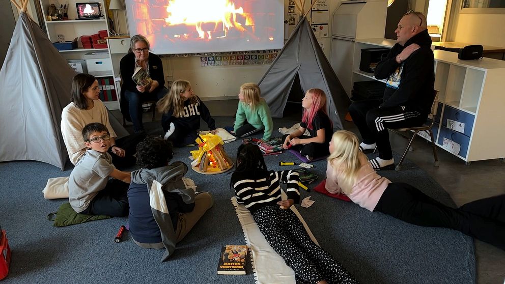 En grupp barn och vuxna samlas i ett rum med tältliknande strukturer. De sitter på golvet omkring en gestaltningsbrand som projiceras på väggen. Några barn har böcker framför sig, medan en vuxen står i närheten och pratar. Atmosfären är avslappnad och lekfull.