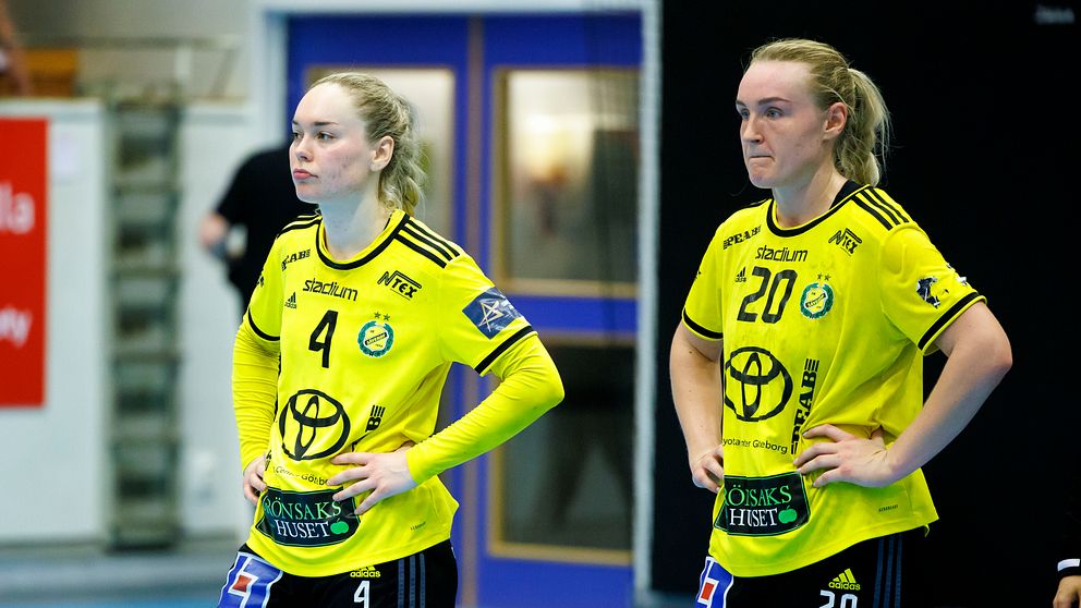Två handbollsspelare står på planen, klädda i gula matchtröjor med svarta detaljer. Den ena spelaren har nummer 4 på bröstet och den andra har nummer 20. De ser fokuserade ut och väntar på spelets fortsättning. Bakgrunden visar en sportarena med blått golv och publikområde.