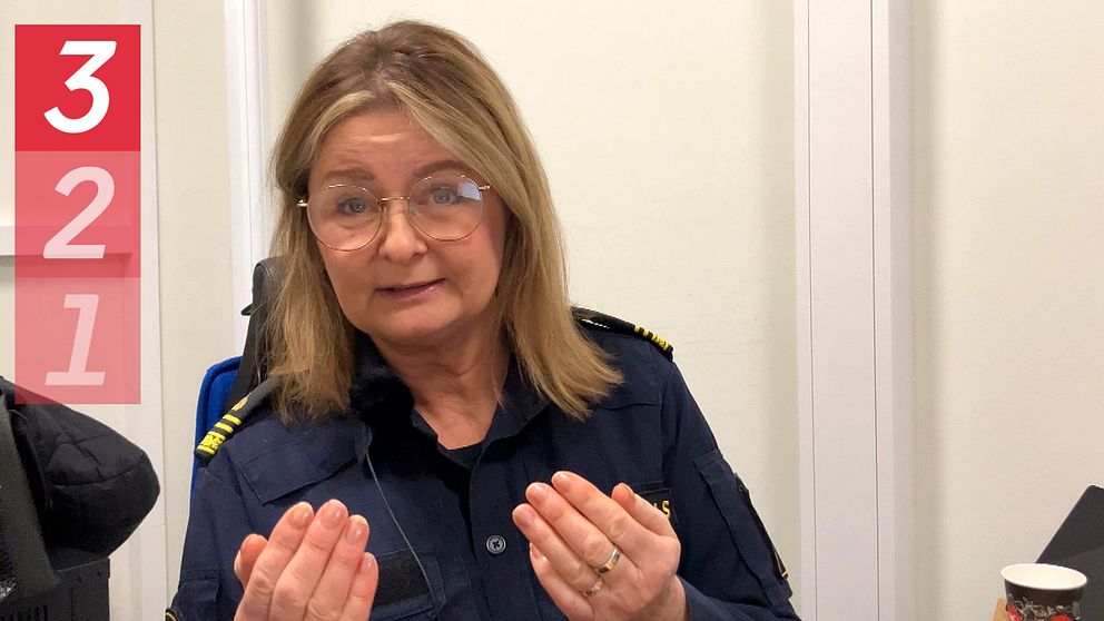 Kvinnlig polis med långt hår och glasögon sitter i förhörsrum och tittar in i kameran. Framåtlutad och gestikulerar med händerna.