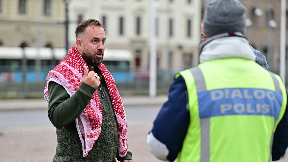 En man står med en hand upphöjd i en diskussion med en polis, som bär en väst med texten ”DIALOG POLIS”. Mannen har en traditionell klädsel med en röd och vit sjal över sin axel. Bakgrunden visar en stadsmiljö med byggnader och en spårvagn i avståndet.