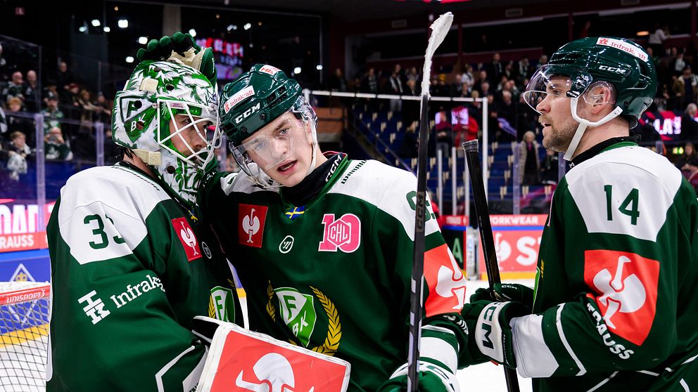 Tre hockeyspelare i gröna och vita dräkter firar en prestation på isen. De är i en jublande omfamning med hockeyutrustning på sig och en publik i bakgrunden. Det är en energisk och glad atmosfär efter en match.
