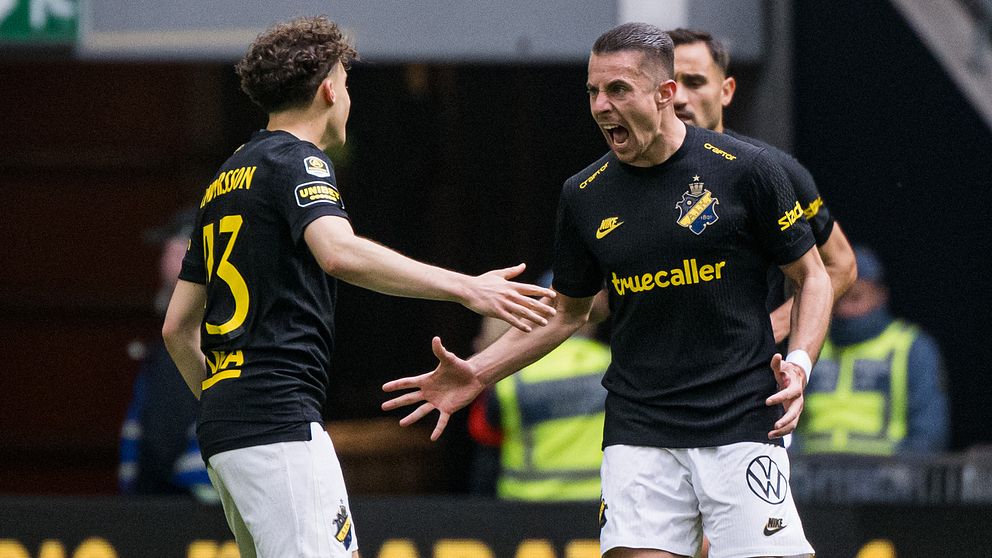 Två AIK-spelare på fotbollsplan med sträckta händer, publik och vakter i bakgrunden.