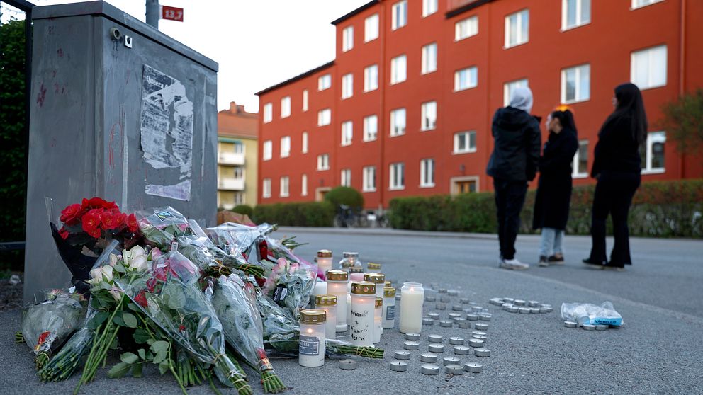 Blommor och tända ljus på trottoar, tre personer samlade i bakgrunden vid rött bostadshus.