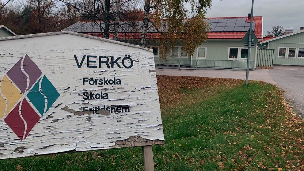 Skylt vid Verkø Förskola, Skola och Fritidhem med en suddig bakgrund av byggnaden och omgivande träd i höst. Skyltens text visar namnet på institutionen och dess verksamheter.