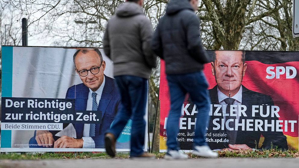 Två personer går förbi affischer med politiska budskap inför ett val i Tyskland. En affisch till vänster tillhör CDU och har texten ”Der Richtige zur richtigen Zeit. Beide Stimmen CDU.” Affischen till höger från SPD har texten ”KANZLER FÜR MEHR SICHERHEIT.” Bakgrunden visar träd utan löv, indicating winter season.