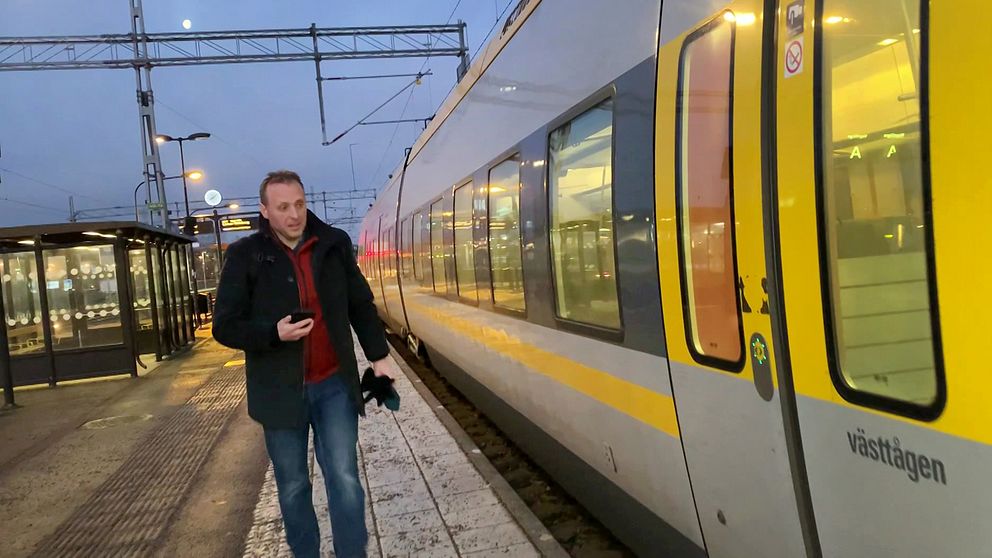 En person går längs en perrong bredvid ett Västtåg som står stilla på stationen. Tåget har gul och grå design och det är skymning utomhus. Plattformen har några lyktstolpar och en väntsal i bakgrunden.