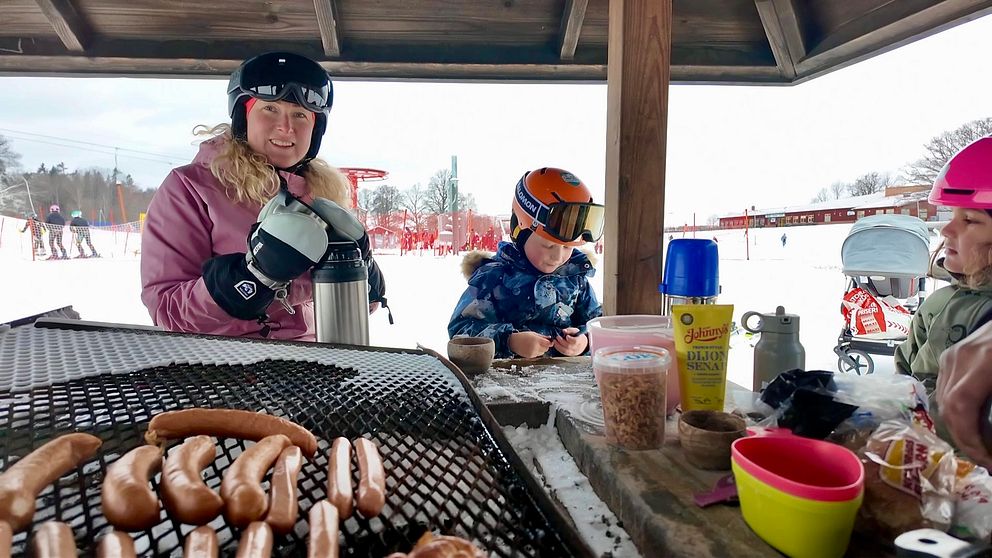 I förgrunden, korvar på grillgaller. Bakom en kvinna i rosa skiddräkt och slalomhjälm