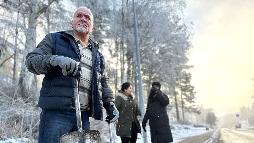 Man som står vid en bilväg med skyffel och krycka i händerna med två kvinnor i bakgrunden.