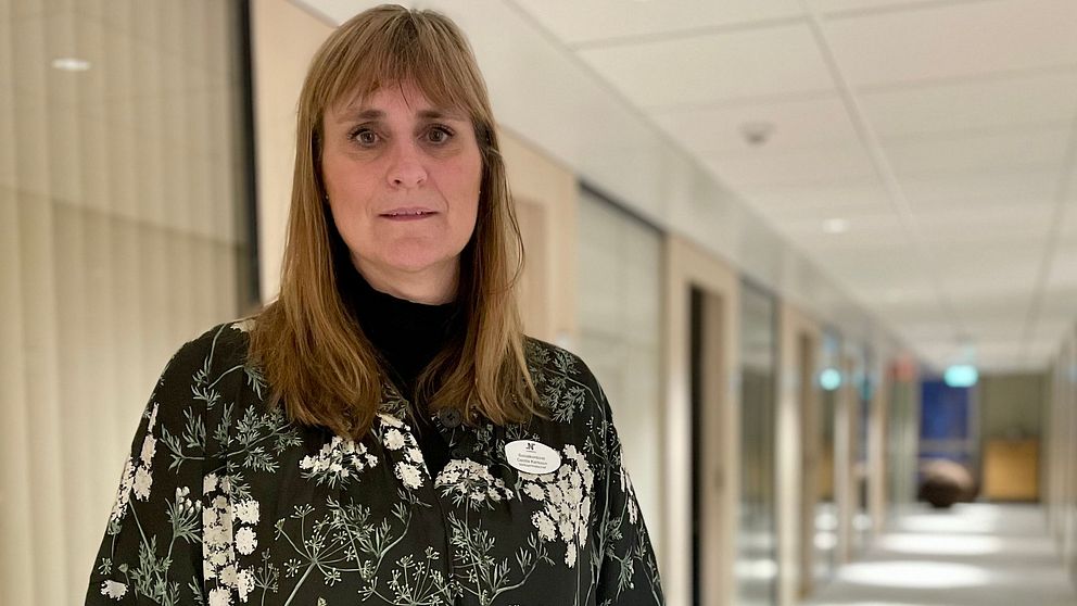 En person står i en korridor med glaspartier längs väggarna. Personen bär en blommig outfit och uttrycker en professionell attityd. Korridoren är belyst av mjukt ljus som kommer från fönstren, och i bakgrunden syns fler kontorsutrymmen.