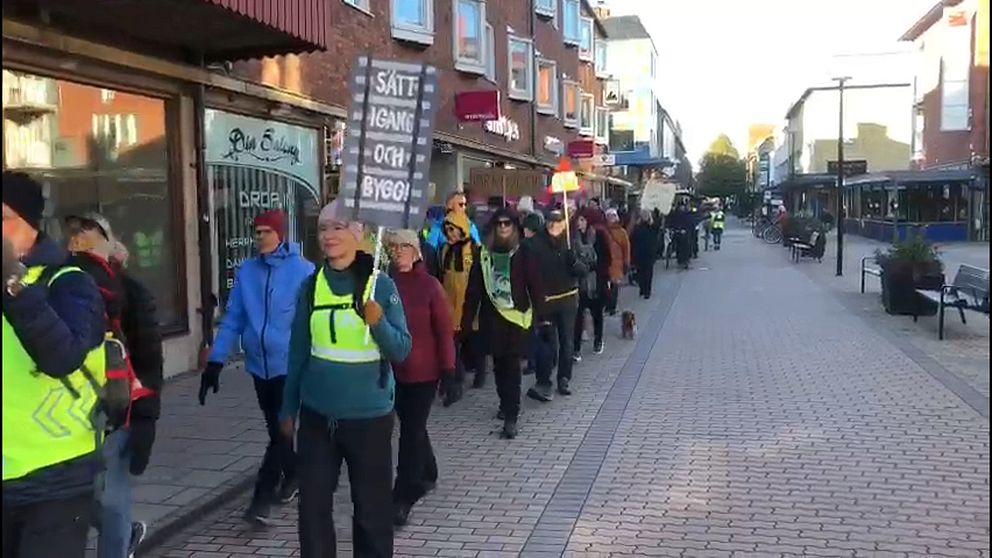 Människor deltar i en demonstration och går längs en gata, bärande på plakat. De flesta har på sig reflexvästar och olika kläder, och de syns representera en grupp med gemensamt syfte. I bakgrunden syns affärslokaler och stadsarkitektur.