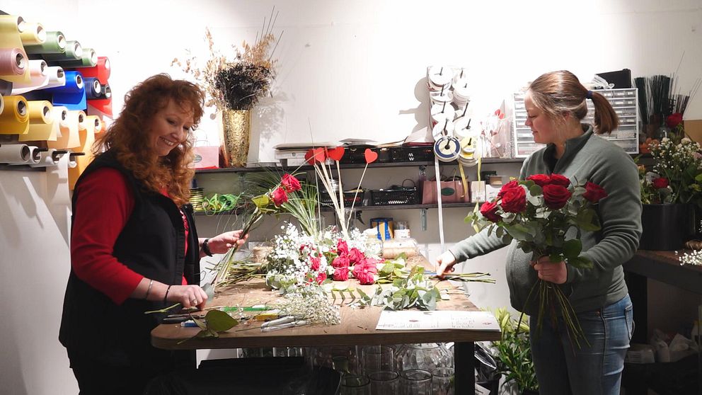 Två kvinnor i en blomsterbutik arrangerar buketter. Den ena kvinnan håller i en ros, medan den andra håller flera blommor. Arbetsytan är fylld med blommor och blad.