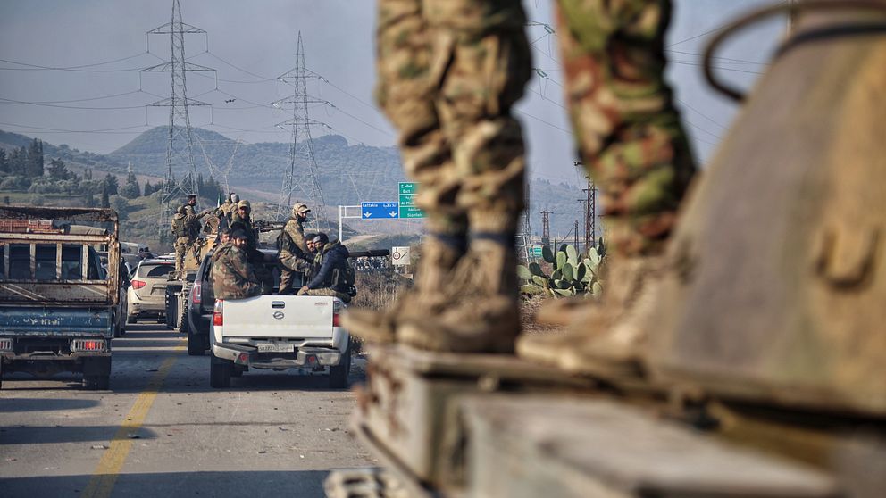 Militär konvoj på väg, soldater på fordon och omgivande landskap med berg i bakgrunden.
