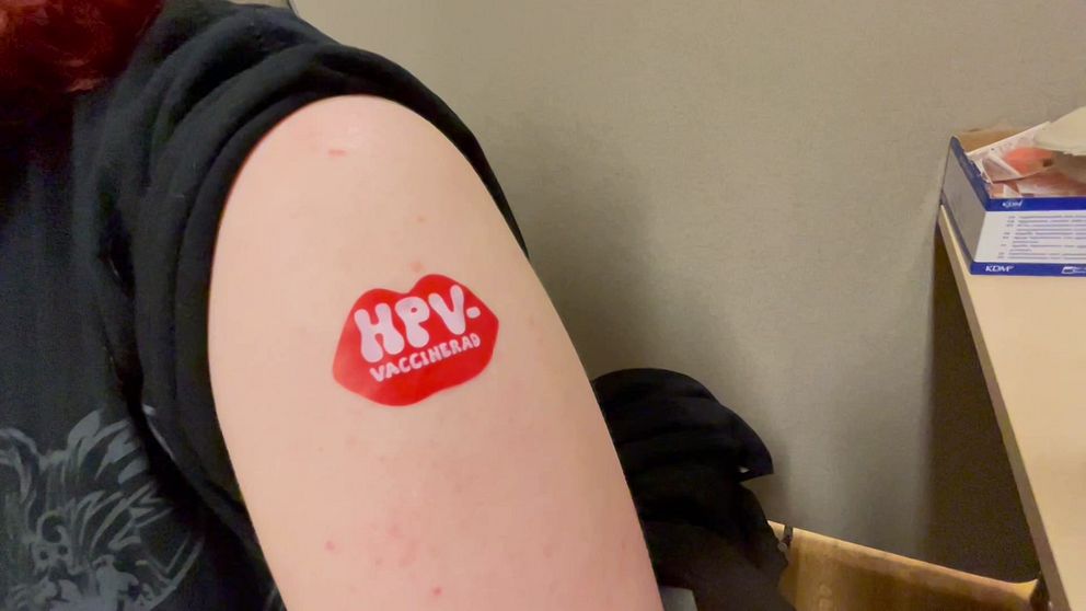 Överarm med klistermärke efter HPV-vaccination. Del av väntrum syns i bakgrunden.