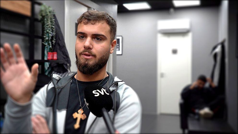 Person intervjuas av SVT inomhus, kors runt halsen, ytterligare person sitter i bakgrunden.